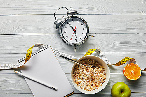 top view of alarm clock, notebook with pencil, breakfast cereal in bowl, apple and orange near measuring tape on wooden white background