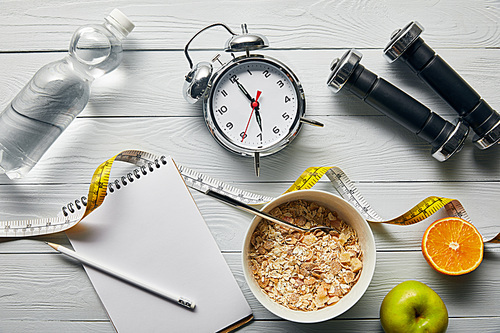 top view of alarm clock, dumbbells, water, notebook with pencil, breakfast cereal in bowl, apple and orange near measuring tape on wooden white background