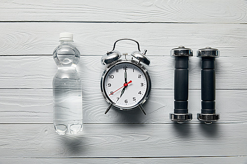 flat lay with silver alarm clock, bottle of water and dumbbells on wooden white background