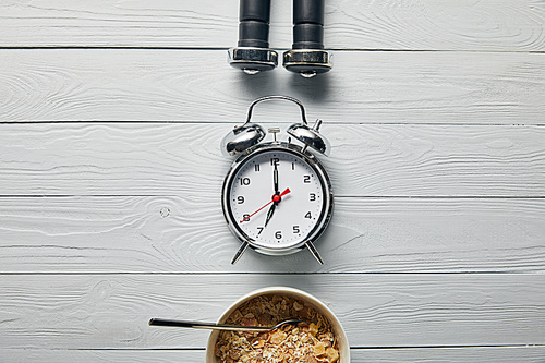 flat lay with silver alarm clock, breakfast cereal in bowl and black dumbbells on wooden white background