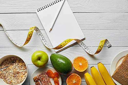 top view of measuring tape, diet food and blank notebook on wooden white background