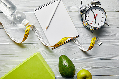top view of notebook and pencil, alarm clock, measuring tape, avocado, apple and water on wooden white background