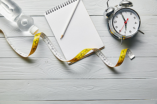 top view of measuring tape, blank notebook, water and alarm clock on wooden white background with copy space