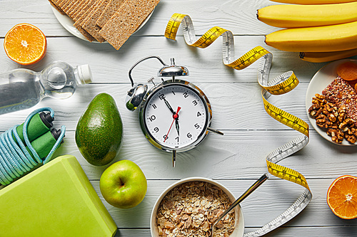 top view of diet food near measuring tape, alarm clock and sport equipment on wooden white background