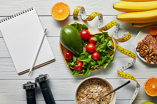 top view of measuring tape, cereal, fruits and vegetables in heart-shaped bowl and dumbbells near blank notebook on wooden white background