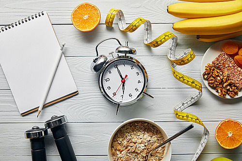 top view of measuring tape, diet food and dumbbells near blank notebook and alarm clock on wooden white background