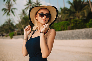 attractive fashionable woman in hat and dress standing on sandy ocean beach