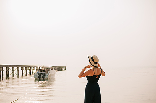 rear view of woman standing in dress and touching hat on beach