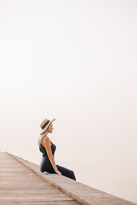 side view of beautiful woman in hat and dress sitting on pier near ocean