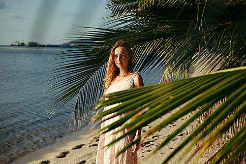 attractive woman standing between palm tree leaves on ocean beach