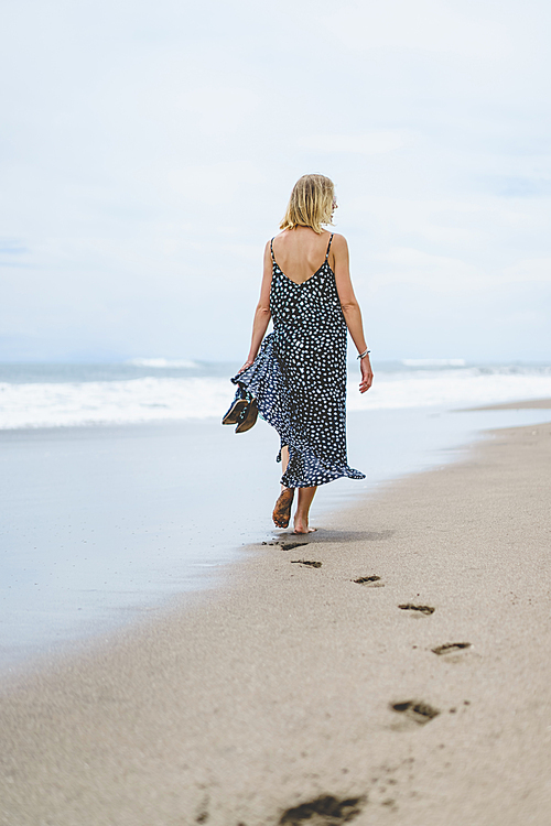 rear view of blonde woman in long dress walking on sandy beach near ocean