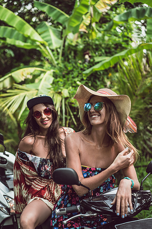 beautiful young female friends in sunglasses sitting on motorcycle in jungles