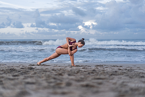 side view of fit young woman practicing yoga in Revolved Crescent Lunge pose (Parivrtta Anjaneyasana) on seashore
