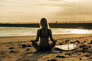 back view of woman meditating on beach at sunset with surfboard near