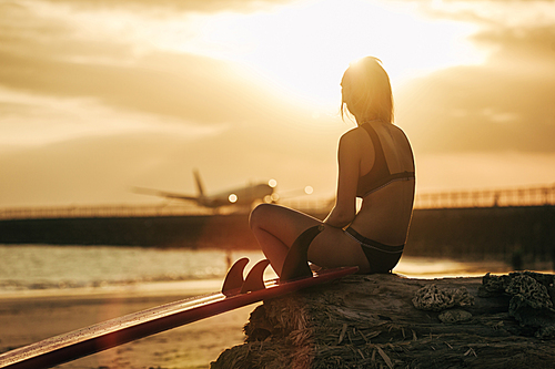 girl sitting on rock with surfboard on beach at sunset with airplane in sky