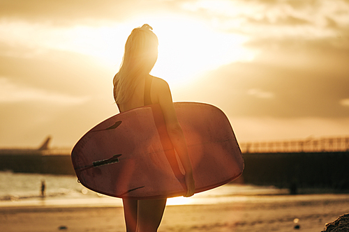 rear view of surfer posing with surfboard on beach at sunset with backlit