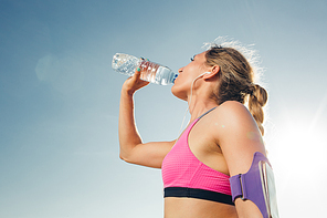 side view of young sportswoman in earphones with smartphone in armband sport case drinking water from bottle against blue sky