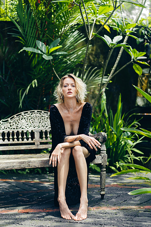 blond woman in black clothing with cigarette in hand resting on bench on terrace, ubud, bali, indonesia