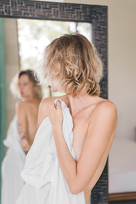 partial view of mirror reflection of blond woman covering body with white bedsheet