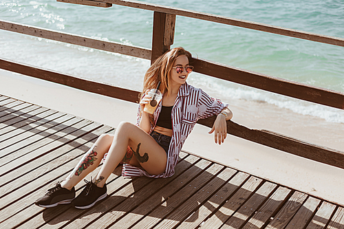 stylish young woman with plastic cup sitting on wooden pier at beach