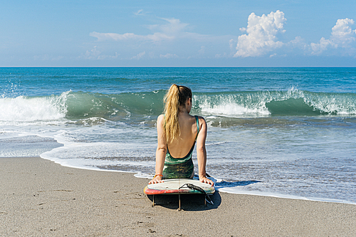 back view of female surfer in swimsuit sitting on surfboard on beach at the sea