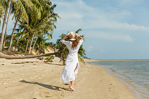 back view of slim girl in white dress walking on tropical beach with palms