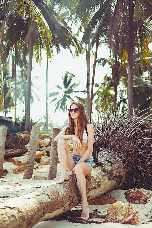 young woman in sunglasses with backpack sitting on fallen palm tree on beach