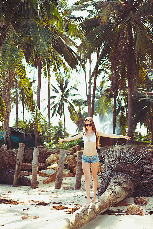 young woman in sunglasses walking on fallen palm tree on beach