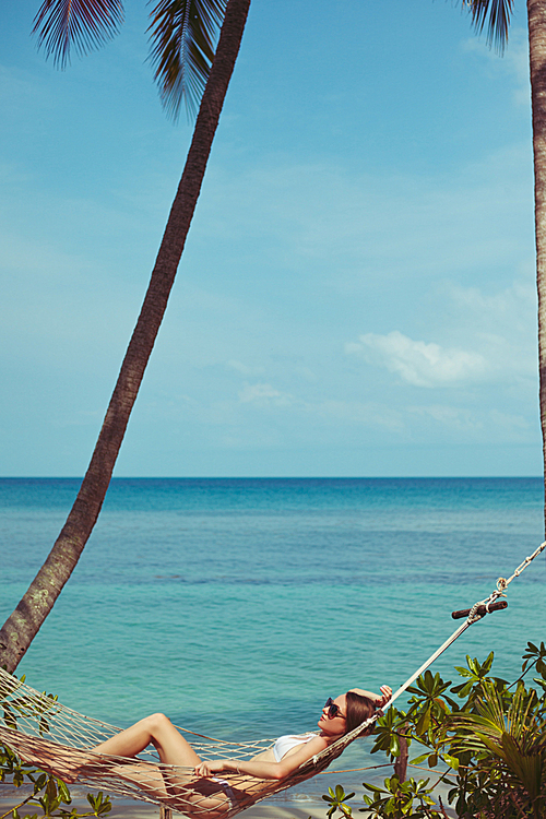 side view of young woman in bikini resting in hammock with ocean on background