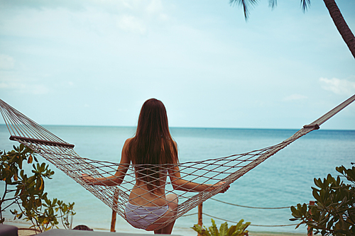 back view of woman in bikini resting in hammock with ocean on background