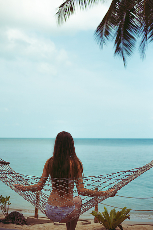 back view of woman in bikini resting in hammock with ocean on background