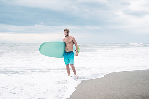 shirtless surfer standing with surfboard and looking at ocean in Bali, Indonesia