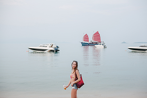 attractive 20대 여성 with cocktail standing on ocean beach with yachts on background