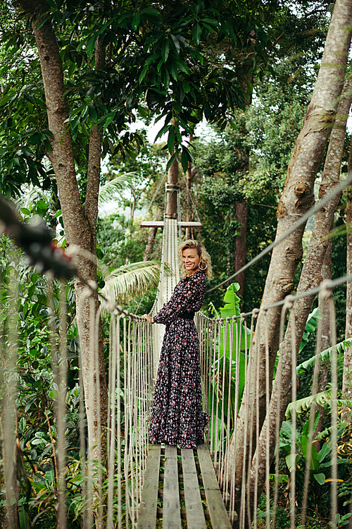 smiling attractive woman standing on footbridge in jungle and looking away