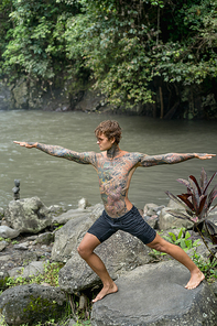 tattooed man practicing yoga on rock with river and green plants on background, Bali, Indonesia