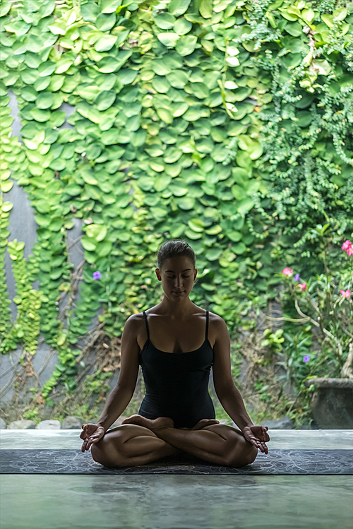 fit young woman practicing yoga in lotus pose (padmasana) with namaste mudra in front of wall covered with green leaves