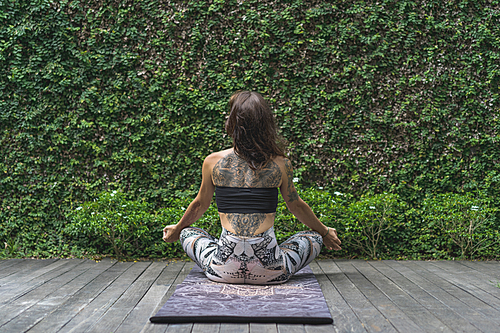 rear view of woman practicing yoga in lotus pose in front of wall covered with green leaves