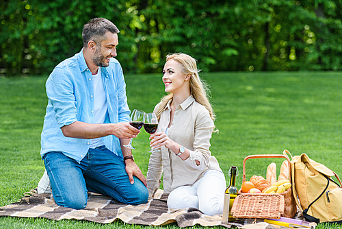 happy couple clinking glasses of wine while sitting together on plaid at picnic