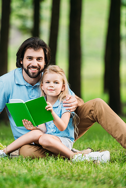 happy father and daughter reading book while sitting on grass at park and 