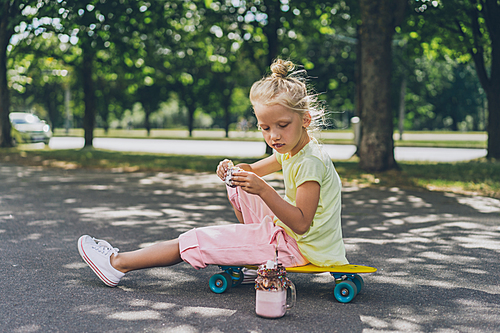 selective focus of adorable kid eating donut from dessert while sitting on skateboard at street