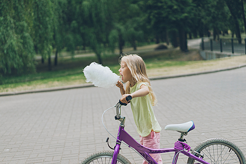 side view of adorable kid with bicycle eating cotton candy in park