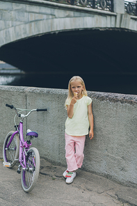 little child eating ice cream while standing near bicycle on street