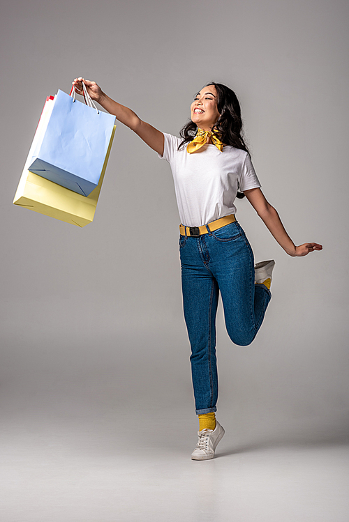 Happy asian woman holding colorful shopping bags in stretched hand on grey