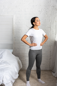 Young asian woman in white t-shirt and grey leggings standing in bedroom holding hands to hips