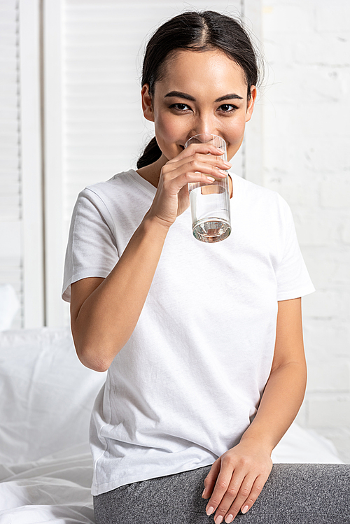 Attractive asian girl in white t-shirt drinking water while sitting on bed