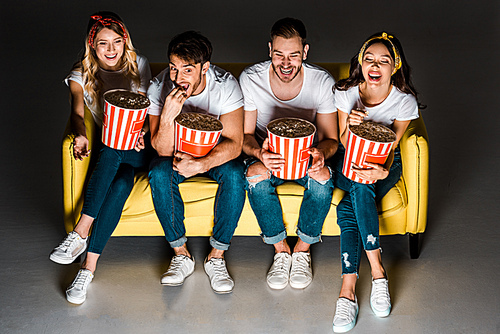 high angle view of happy young friends sitting on couch and eating popcorn from boxes on grey