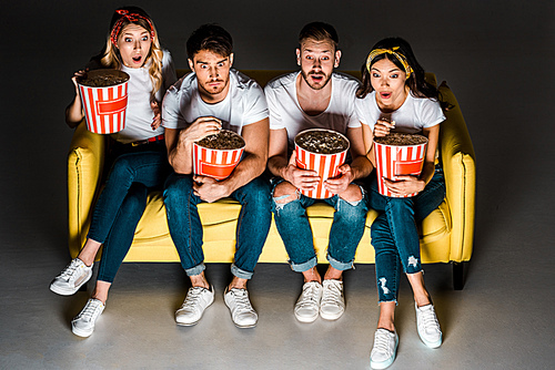 high angle view of shocked young friends holding popcorn boxes and watching tv while sitting together on sofa on grey