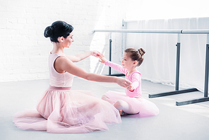 side view of ballet teacher and little student in pink tutu skirts sitting and holding hands while training in ballet school