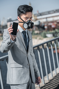 asian businessman in gas mask showing smartphone on bridge, air pollution concept
