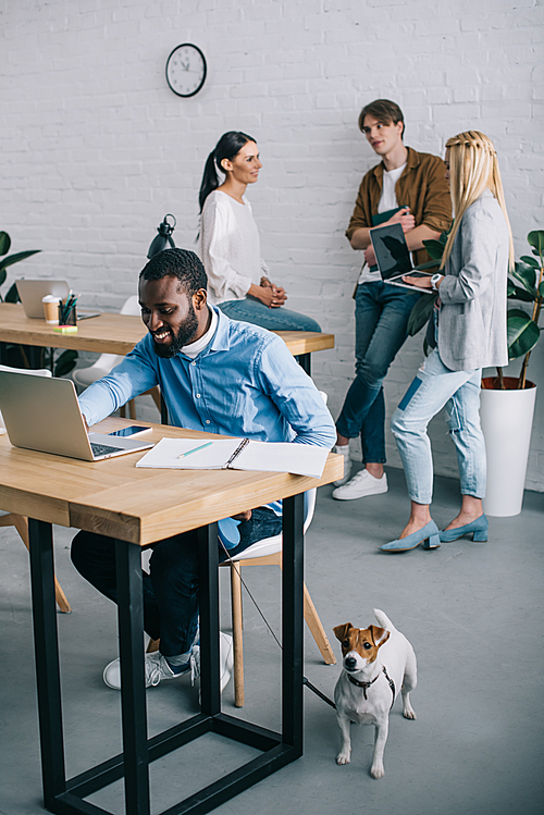 smiling african american businessman working on laptop and holding dog on leash and coworkers having meeting behind
